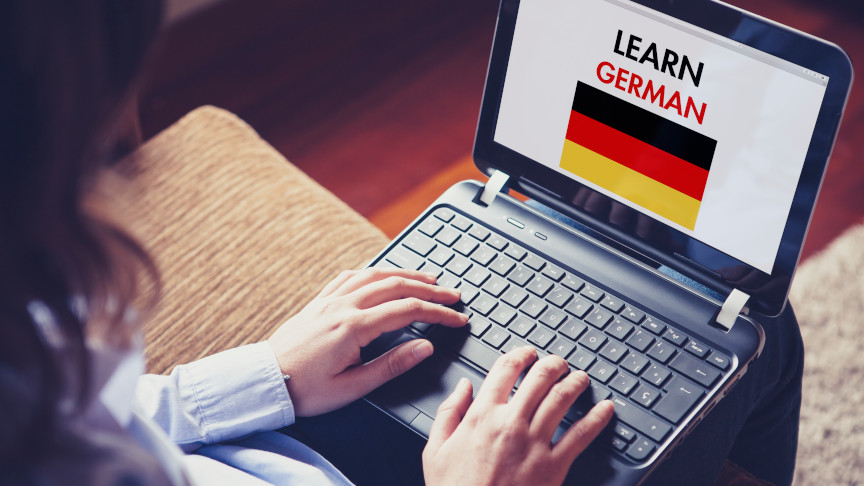 Woman sitting at home with a laptop on the knees and a website to learn German on the screen.
